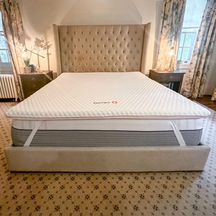 A mattress with a Dormeo topper rests on a bed frame in a bedroom with nightstands, lamps, and floral curtains.