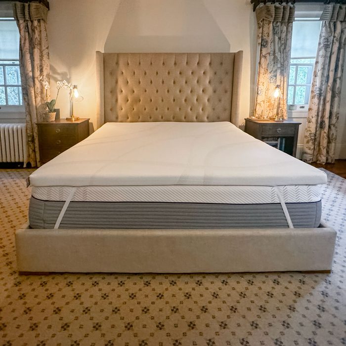 A large bed with a beige tufted headboard sits in a bedroom, flanked by nightstands with lamps and ornate floral curtains.