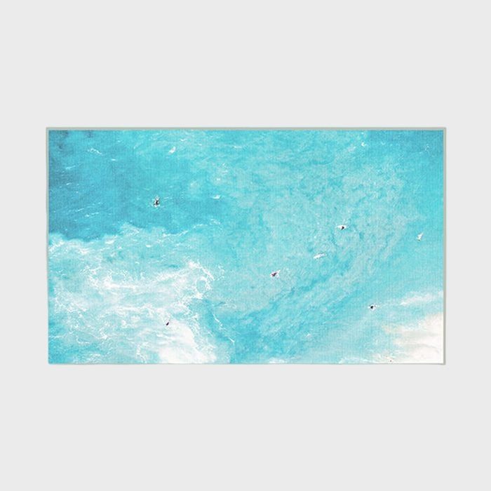 Surfers float on bright turquoise waves, viewed from above; the sea shimmers with white foam under a clear, expansive sky.