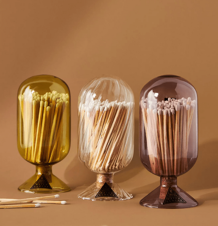 Glass jars hold wooden matches, standing upright in a gradient of amber, clear, and smoky hues, against a warm brown backdrop.