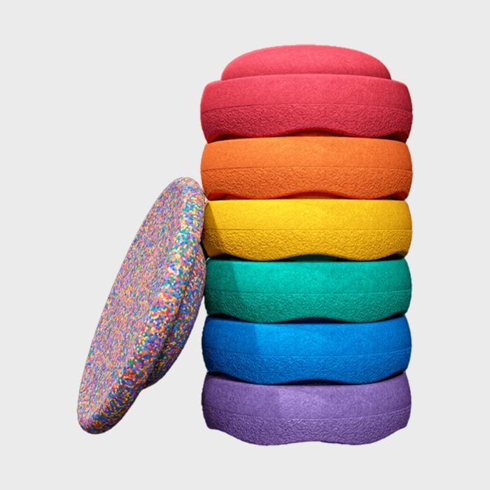 Stacked colorful round cushions display vibrant hues; red, orange, yellow, green, blue, and purple, with one speckled cushion leaning against them, isolated on gray background.