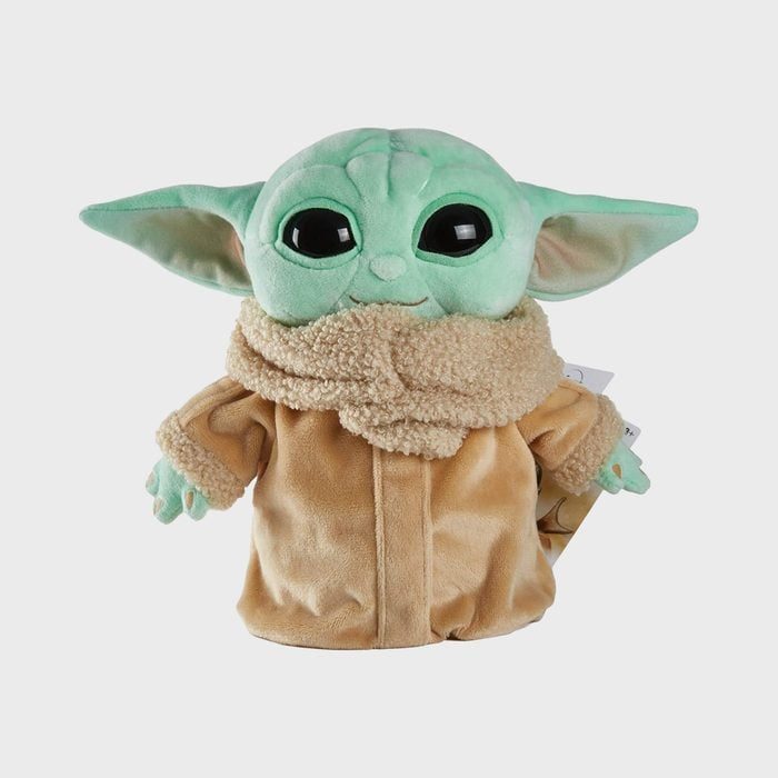 A green plush toy stands wearing a brown robe and scarf, displaying large eyes and prominent ears against a plain background.