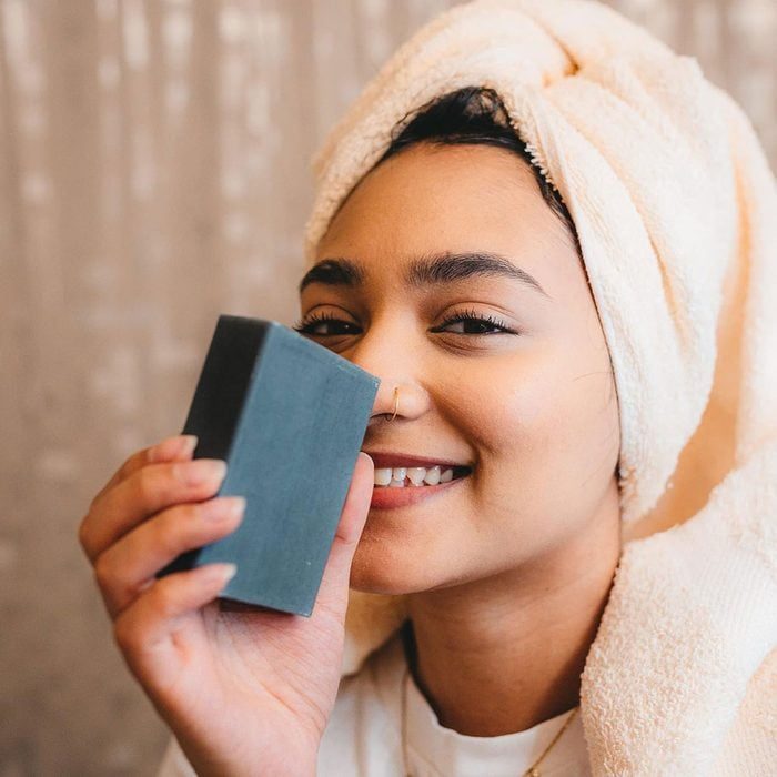 A woman with a towel on head holds black soap, smiling in a cozy bathroom setting.