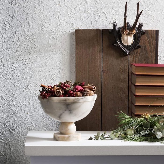 A marble bowl holds dried potpourri, sitting on a white shelf. A wooden wall with antlers decor is behind, alongside a stack of books.