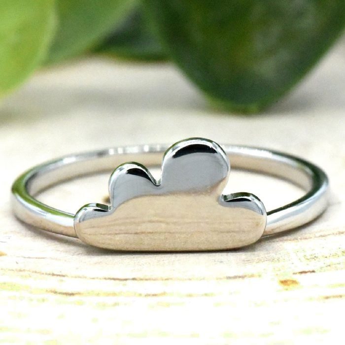 Silver ring features a cloud-shaped design, resting on a wooden surface with blurred green leaves in the background.