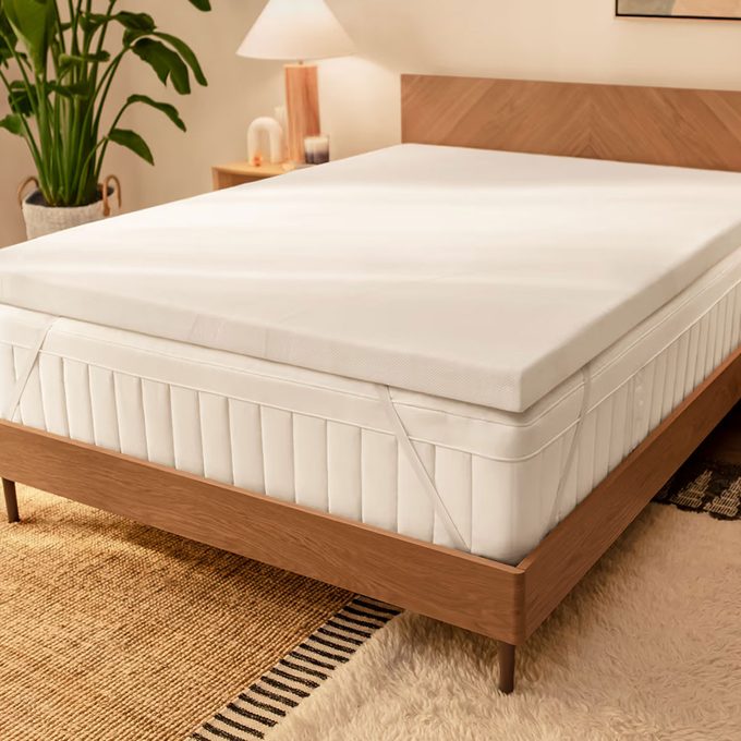 Bed with clean sheets sits in a cozy room, surrounded by a wooden headboard, bedside lamp, plant, and textured rugs.