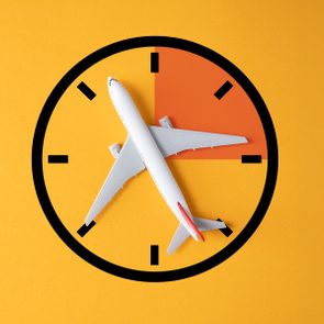 Airplane positioned diagonally across a clock face on a yellow background. The clock has a quarter orange segment, indicating time-related travel theme.
