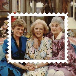 Three women smiling, sitting closely on a colorful floral couch, framed like a stamp in a cozy, warmly lit living room.