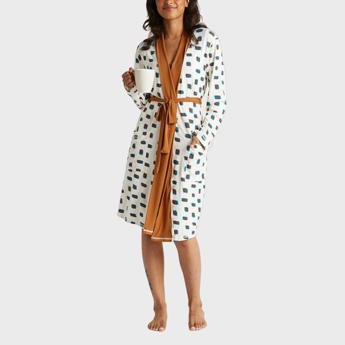 Person holds a mug, wearing a white robe with blue squares and orange trim, standing barefoot against a plain background.
