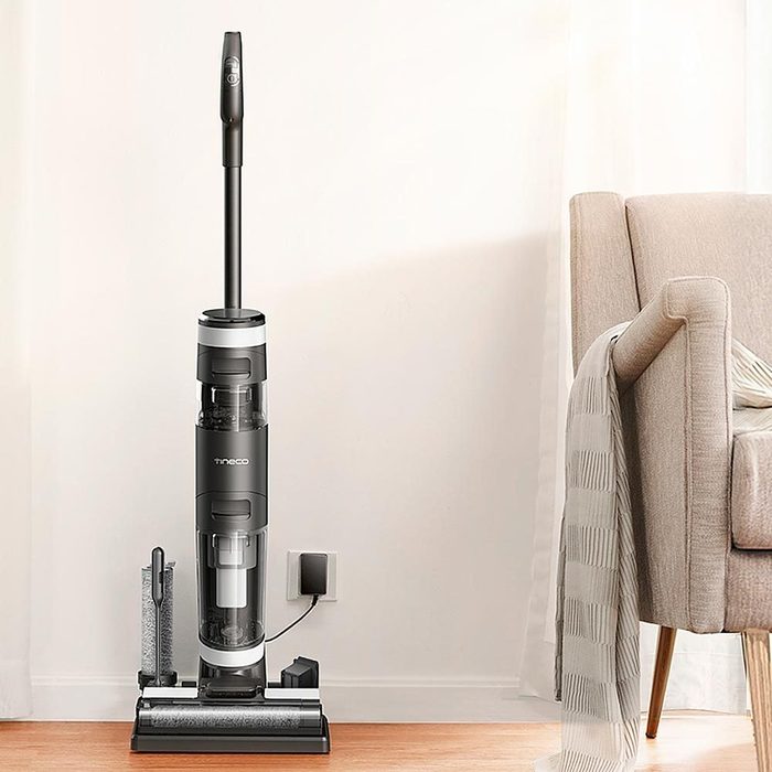Vacuum cleaner stands upright, charging on a wooden floor near an armchair, next to a wall outlet.