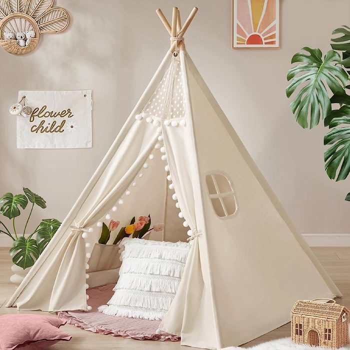 A cream-colored teepee houses pillows and tulips, surrounded by plants in a cozy room. A banner reads 