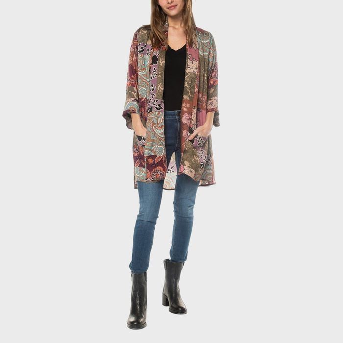 Woman stands wearing a colorful, patterned coat over a black top and jeans, with hands in pockets; black boots complete the outfit.