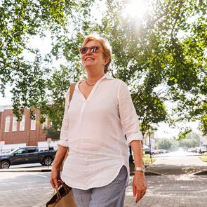 A person wearing sunglasses walks confidently while smiling; the setting is a sunny day on a tree-lined street with buildings and parked cars.