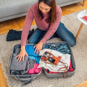 A person packs clothes and a camera into a suitcase on the floor, near a white table with drinks and documents.