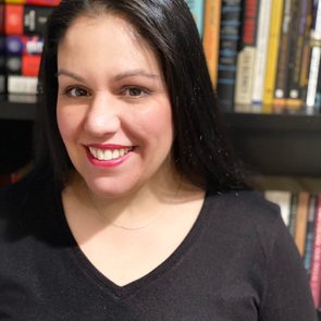 Person smiling, wearing a black top, standing in front of a full bookshelf.
