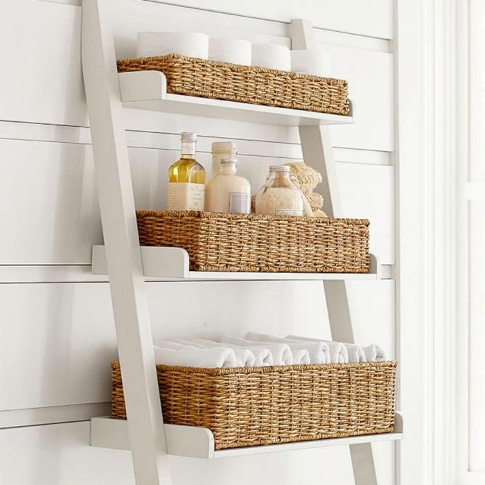 Wicker baskets rest on a white ladder shelf, holding toiletries and towels, against a paneled wall.