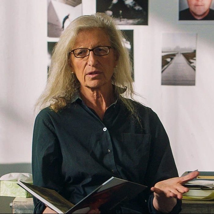 A person holds an open book, gesturing, with photographs displayed on the wall behind them.