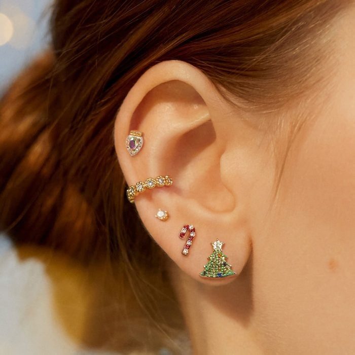 Ear adorned with various earrings: heart, hoop, pearl, candy cane, Christmas tree. Set against a warm, softly blurred background.