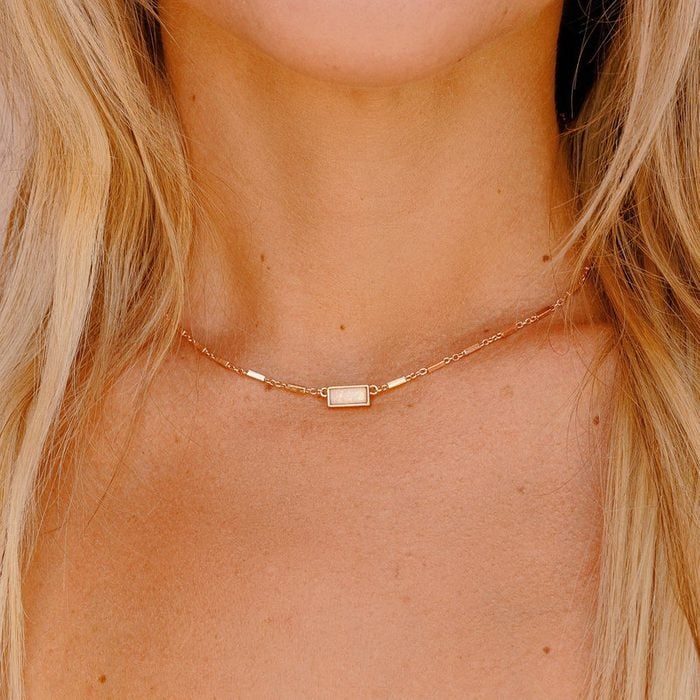 Necklace rests on a woman's neck, featuring a rectangular pendant, surrounded by blonde hair, against a neutral background.