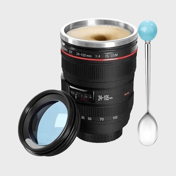 Coffee cup shaped like a camera lens holds coffee. Lid and spoon with blue handle beside it, against a plain background. Text: 
