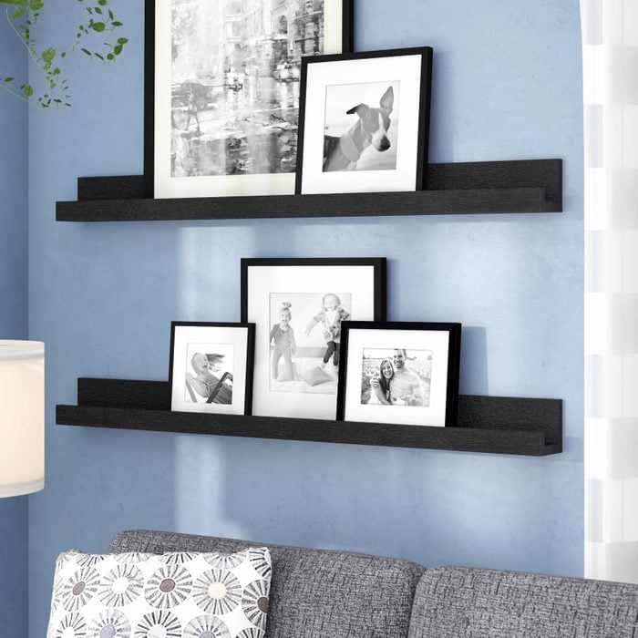 Framed photographs sit on two black wall shelves against a light blue wall. A gray sofa with patterned cushions is visible below.