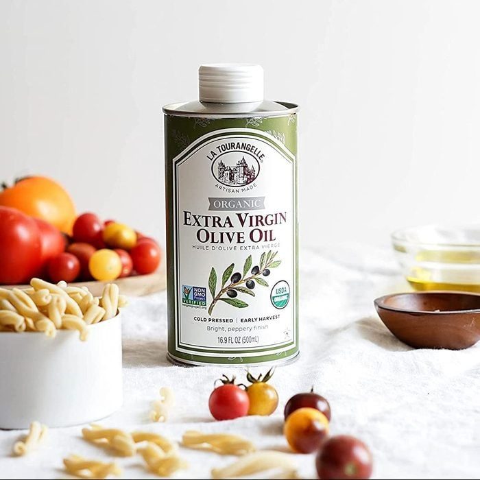 Olive oil tin stands on a white cloth, surrounded by tomatoes, pasta, and a bowl, creating a fresh cooking theme. Text: 