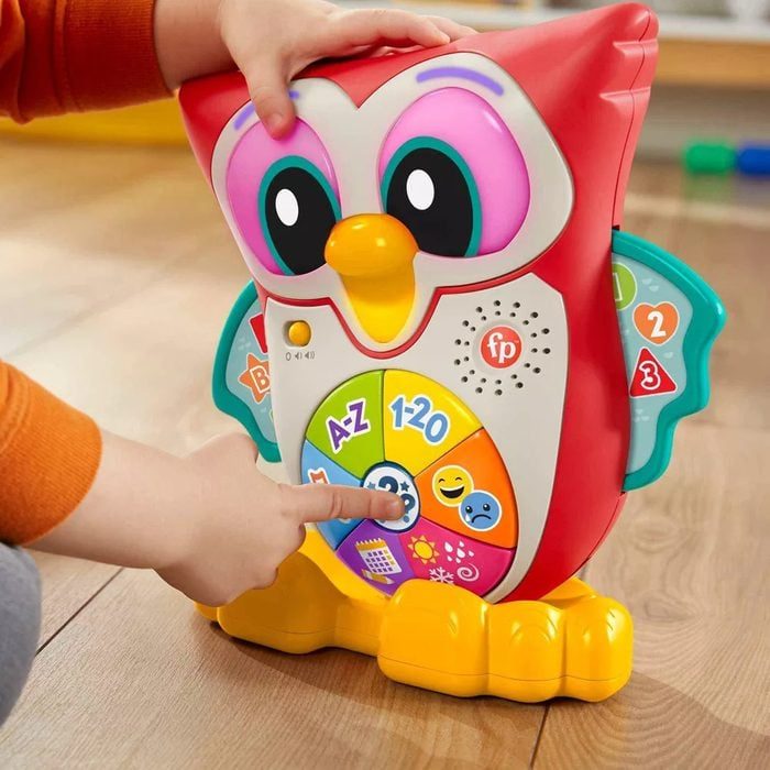 Toy owl displays colorful buttons; child presses, activating sound functions. Located on wooden floor, emphasizing playful learning with letters, numbers, and emojis.