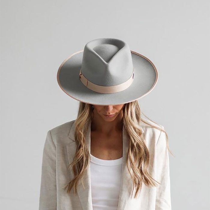 A woman wears a gray wide-brimmed hat, standing with her head down, indoors against a plain light background, dressed in a light-colored blazer.