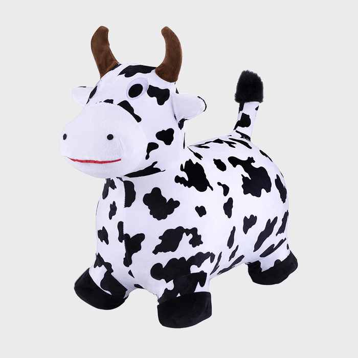 A black-and-white spotted cow toy stands, featuring brown horns and a small tail, set against a plain, light gray background.