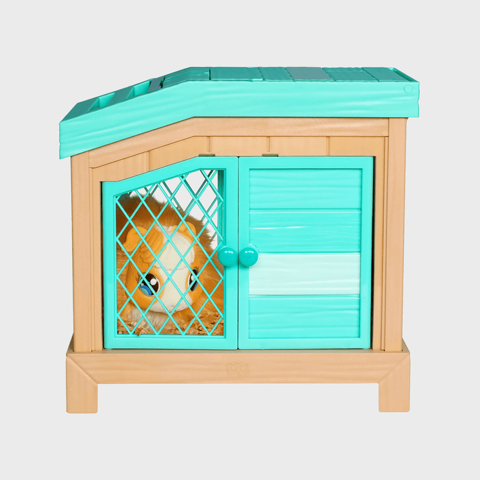 Toy hamster sitting inside a turquoise and tan plastic cage, looking through diamond-patterned bars. The cage has a slanted roof and two doors.