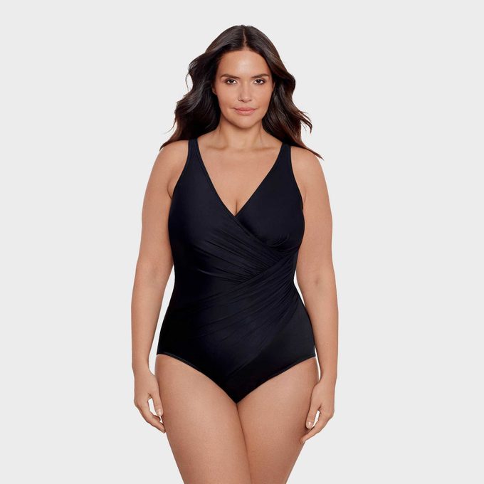 A person stands wearing a black, one-piece swimsuit against a plain white background, looking directly at the camera.
