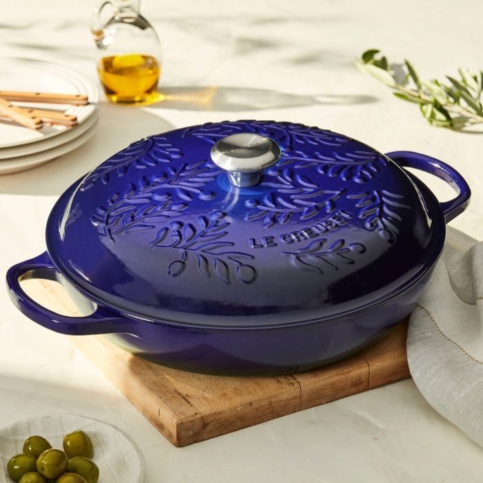 Blue cast iron pot sits on wooden board, surrounded by olive oil bottle, plates, and olives.