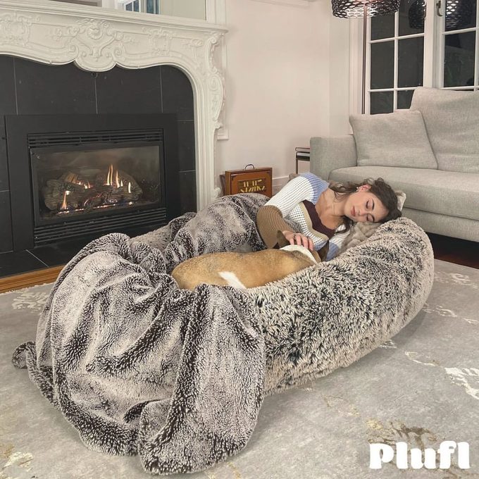 Person sleeps with dog on a large, fuzzy bed in front of a fireplace, next to a couch. Text reads â€œPlufl.