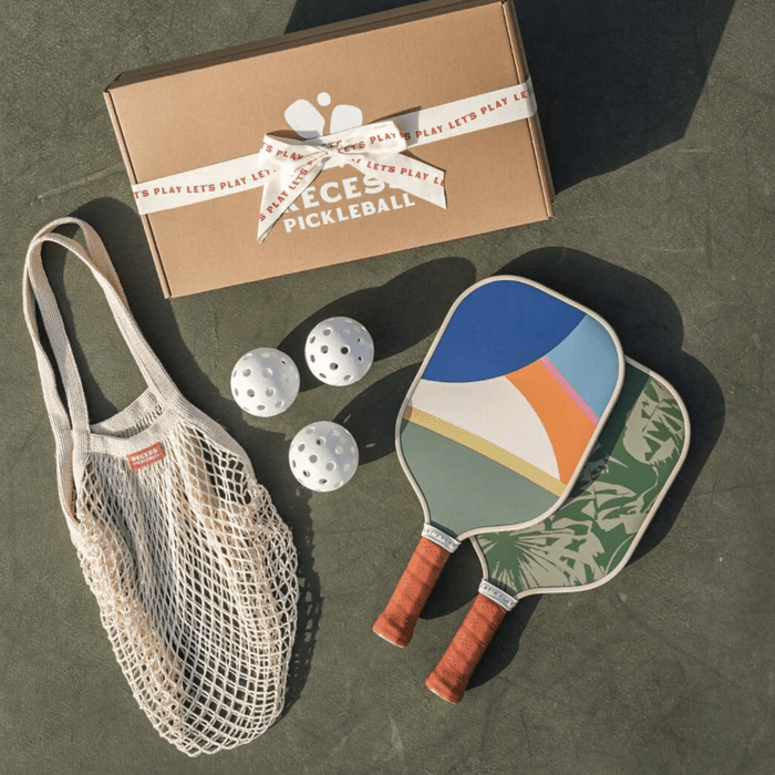 Paddles lie on the ground with pickleball balls nearby, alongside a netted bag and a box labeled 