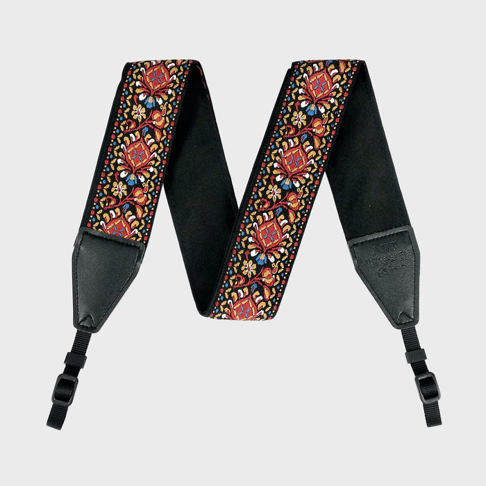 A colorful, embroidered guitar strap displays intricate floral designs on a light grey background, featuring leather ends and adjustable black plastic buckles.