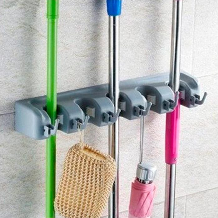 Wall-mounted organizer holds mops and brooms with grips, against a tiled wall. Additional hooks hold a mesh bag and an umbrella.