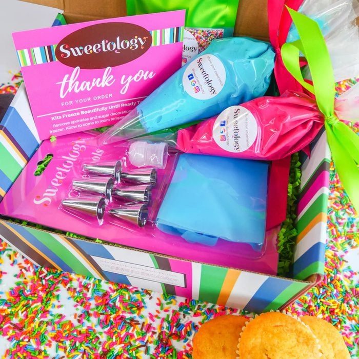 Box contains cake decorating supplies, including icing bags and nozzles. It rests among colorful sprinkles. 