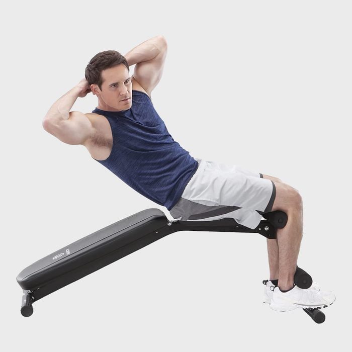 Man exercises on incline bench, performing a sit-up. He wears a blue tank top and white shorts, in a minimal, indoor fitness setting.