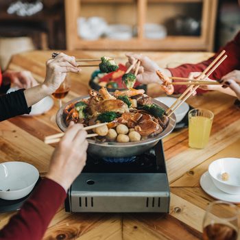 People are cooking and eating food with chopsticks from a shared hot pot on a wooden table, surrounded by drinks and plates.