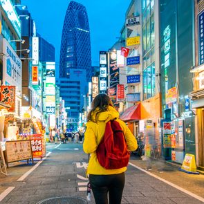 Person walks down a neon-lit street, wearing a yellow jacket and red backpack. Tall buildings and vibrant signs create a bustling urban atmosphere.