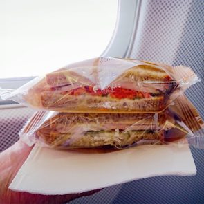 Wrapped sandwiches rest on a napkin in a hand, near an airplane window.