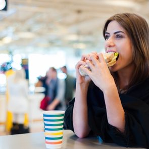 A person eats a sandwich at a table next to a colorful cup in a busy indoor public space.