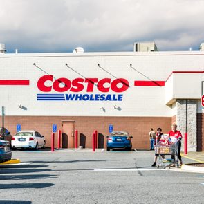 Costco shoppers push carts in a sunny parking lot, with cars and a stop sign nearby. The building is adorned with the 