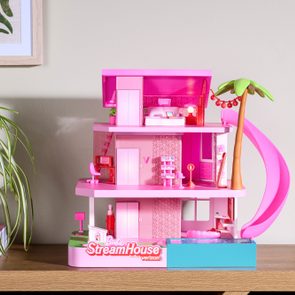 Dollhouse featuring vibrant pink furniture, situated on a shelf near a plant. 