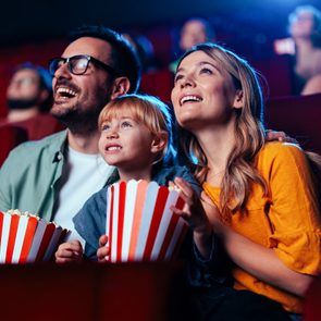 A family watches a movie, smiling and holding popcorn, in a dimly lit theater with red seats.