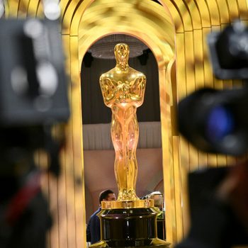Oscar Academy Award statuette beneath a golden arch, with cameras pointed at it in the foreground