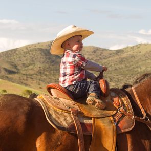 Baby in a tan cowboy hat and flannel shirt riding a horse with mountains in the background