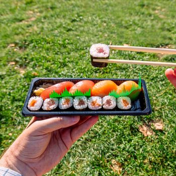 Hand holding a box of takeout sushi in a grassy park, with green plastic grass separating the pieces, and chopsticks holding a piece of sushi above the box