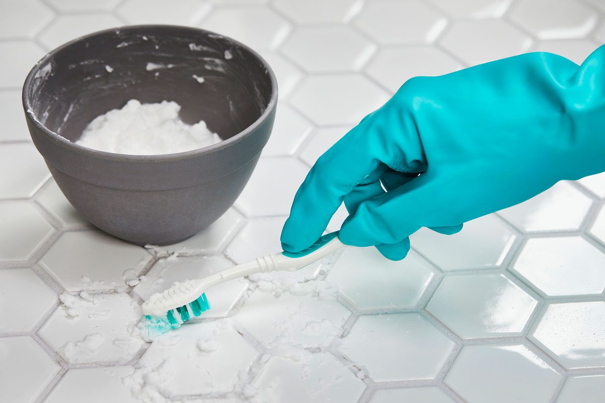 Gloved hand using a toothbrush to scrub baking soda mixture onto grout lines on a white tiled floor