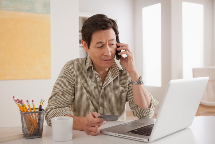 man talking on the phone while holding a credit card and looking at laptop screen